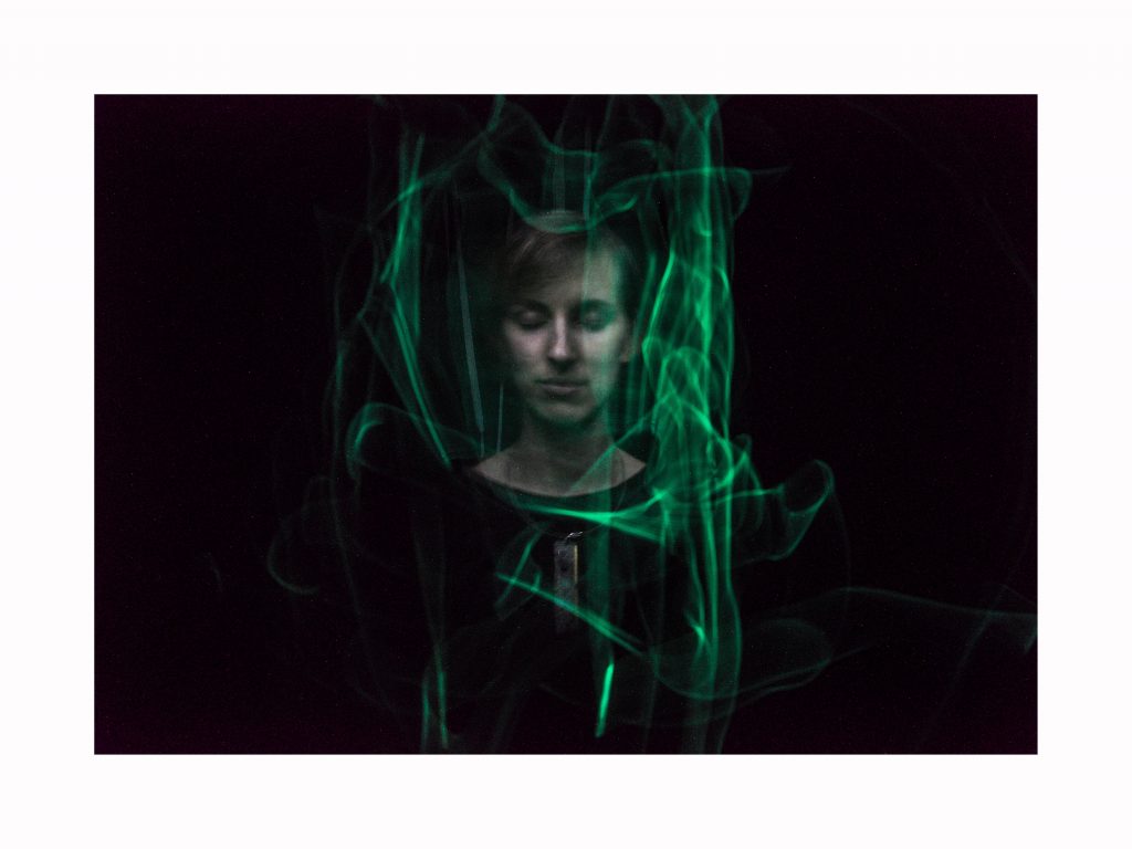 In der Bildmitte sehen wir eine Person vor schwarzem Hintergrund, wir sehen Kopf und Oberkörper. Das Gesicht ist gleichmäßig beleuchtet, die Augen sind geschlossen. Das offenbar kurze glatte blonde Haar fällt nach links über die Stirn. Der Hals ist bis zu dem runden Ausschnitt des schwarzen Pullis zu sehen. Um den Hals trägt die Person einen rechteckigen Anhänger an einem Metalldraht. Um das Gesicht herum tanzen grüne Lichtspuren. Schwächere davon auch direkt vor dem Gesicht. Die Lichtspuren bilden so etwas wie einen eckigen Rahmen um das Gesicht. Die Konturen sind weich, wie gemalt. Das weiche Licht lässt die Haut blaß und fast grau erscheinen. Das Gesicht ist jung und nicht eindeutig männlich oder weiblich. Die Form ist oval, der geschlossene Mund ist entspannt und wie von einem Lächeln umspielt. Die Brauen bilden einen perfekten Bogen über den weit auseinander stehenden geschlossenen Augen. Sie erinnern an Ikonenmalerei in ihrer Gleichmäßigkeit. Die gerade Nase und der genau waagrechte Mund verstärken den Eindruck. Der Rest des Körpers ist nicht zu erkennen und verschmilzt mit dem Hintergrund. Die grünen Lichtspuren verlaufen rings um den Kopf. Links tanzen sie etwa in Entfernung der Breite des Gesichts senkrecht in der Luft. Sie bilden eine kleine Spitze, die nach links zeigt und setzen sich parallel nach hinten fort. Rechts sind sie länger und kräftiger, reichen fast zum unteren Bildrand und bilden mehrere kleine Schleifen direkt neben der Wange. Einige wagen sich bis vor das Gesicht und verschleiern es geheimnisvoll. Oberhalb des Gesichts vereinigen sie sich in einer einzelnen grünen Schleife quer über dem Kopf. Unterhalb laufen sie quer am Auschnitt des Pullis entlang und betonen das längliche Schmuckstück, das entlang des Brustbeins getragen wird. Das Material dieses Schmuckes könnte Metall sein, es schimmert bläulich. Es wird durch vier einzelne grüne Spuren in einem Quadrat eingerahmt. Die gesamten Spuren in ihrer Verteilung erzeugen einen erstaunlichen räumlichen Effekt: sie bilden wirklich einen sichtbaren Schleier um den Kopf der Person. Die unruhigen grünen Lichtstreifen bilden dabei einen starken Stimmungskontrast zu dem in sich ruhenden entspannten Gesicht. Durch diesen Schleier wirkt die Person sehr entrückt, wie aus der Welt gefallen oder gerade aus dem All gebeamt. Beschrieben von Katrin Heidorn